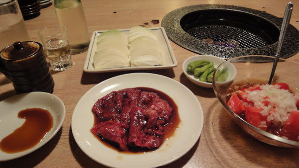 plates of beef, onion, and peppers with a tomato salad and a drink on a table near a grilling brazier